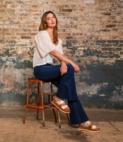 A woman wearing the Bed|Stü Vext brown platform slide sandals, featuring a leather-wrapped sole, sits on a stool against a worn brick wall in a white blouse and blue jeans, looking at the camera.