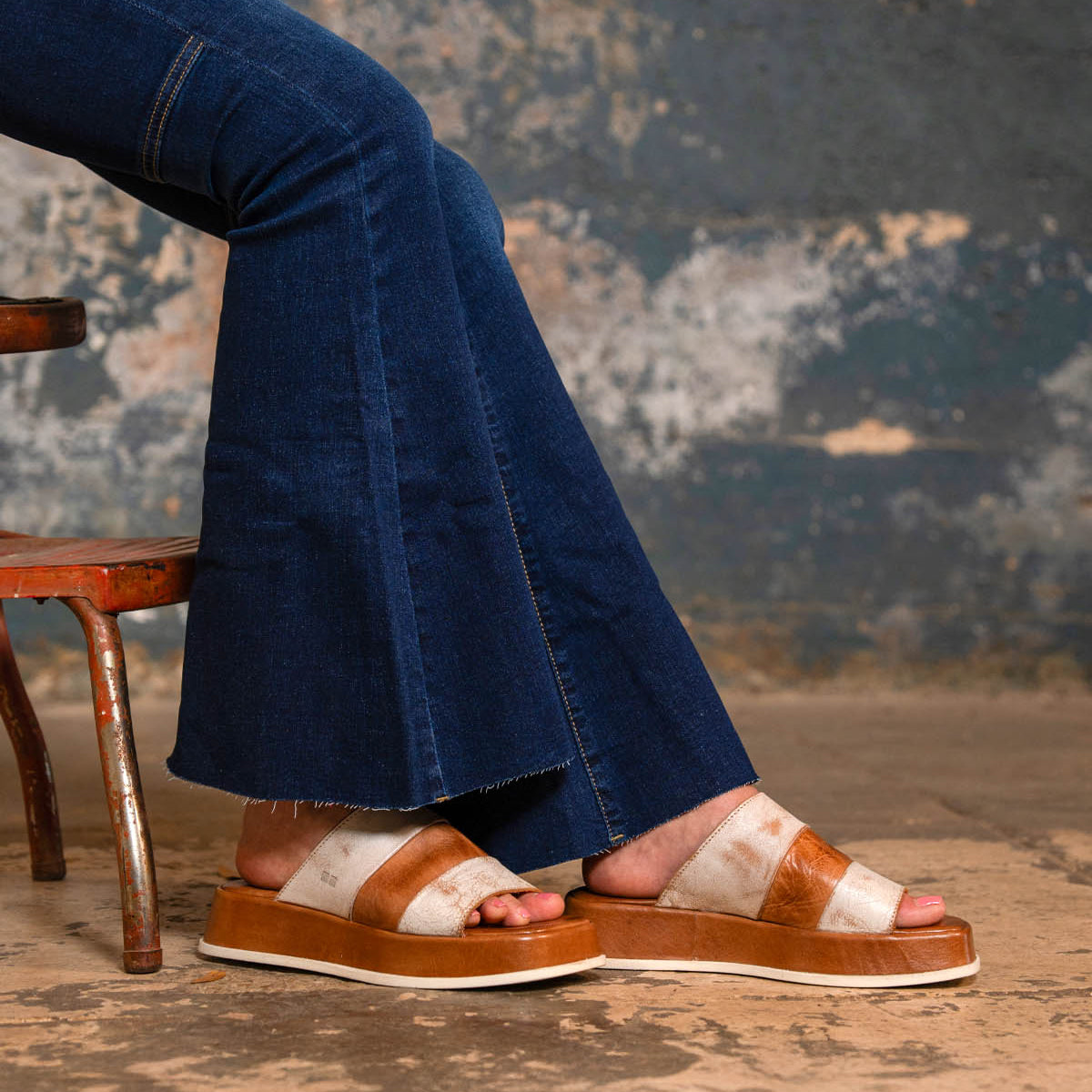 A person wearing flared blue jeans and the Bed|Stü Vext tan-and-white square-toe platform slide sandals with a leather-wrapped sole sits on a distressed chair against a weathered wall.