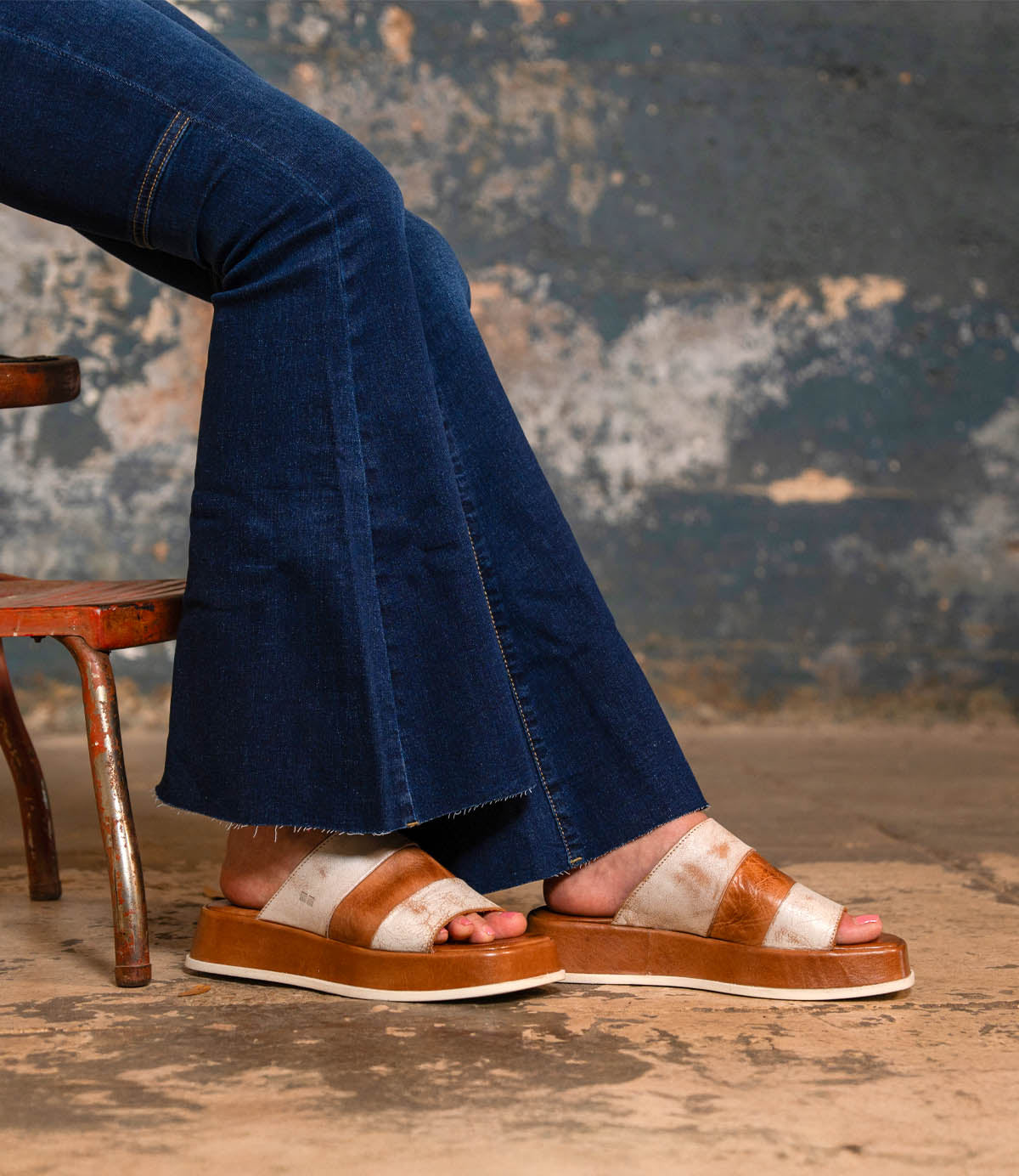 A person wearing flared blue jeans and the Bed|Stü Vext tan-and-white square-toe platform slide sandals with a leather-wrapped sole sits on a distressed chair against a weathered wall.