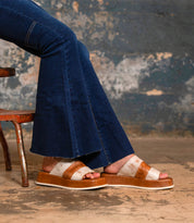 A person wearing flared blue jeans and the Bed|Stü Vext tan-and-white square-toe platform slide sandals with a leather-wrapped sole sits on a distressed chair against a weathered wall.