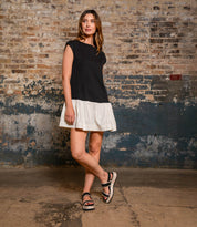 A woman stands indoors against a textured brick wall, wearing a black and white dress with a ruffled hem and chic Bed|Stü Vice platform sandals featuring an adjustable ankle strap.