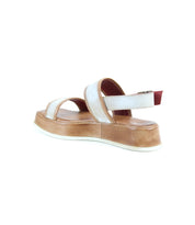 The Bed|Stü Vice features a single strappy design with white straps, a tan platform sole, and a red-accented adjustable ankle strap, showcased against a white background.