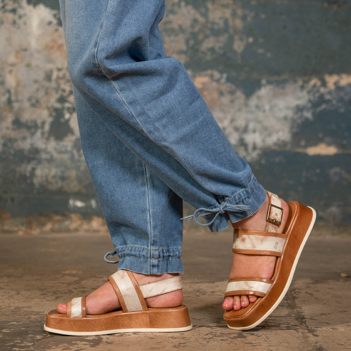 A person wearing Bed|Stü Vice light brown strappy platform sandals with an adjustable ankle strap and blue cuffed jeans stands on a rough concrete floor, with a textured, worn wall in the background.