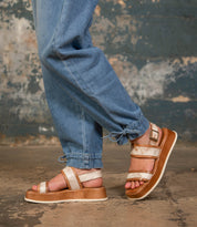 A person wearing Bed|Stü Vice light brown strappy platform sandals with an adjustable ankle strap and blue cuffed jeans stands on a rough concrete floor, with a textured, worn wall in the background.
