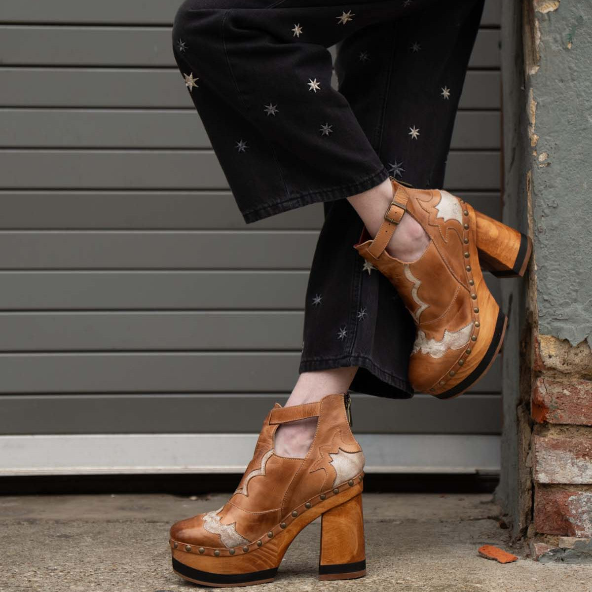 A person wearing black pants with star designs and vintage-inspired Bed|Stü Viretta shoes, featuring a handcrafted wooden platform, stands with one leg crossed in front of a garage door and brick wall.
