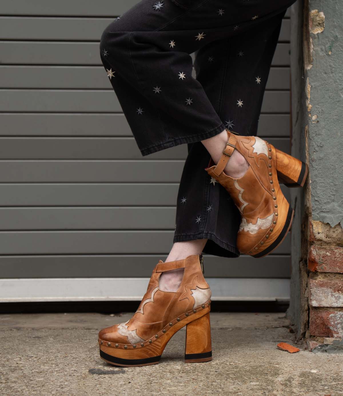 A person wearing black pants with star designs and vintage-inspired Bed|Stü Viretta shoes, featuring a handcrafted wooden platform, stands with one leg crossed in front of a garage door and brick wall.