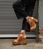 A person wearing black pants with star designs and vintage-inspired Bed|Stü Viretta shoes, featuring a handcrafted wooden platform, stands with one leg crossed in front of a garage door and brick wall.