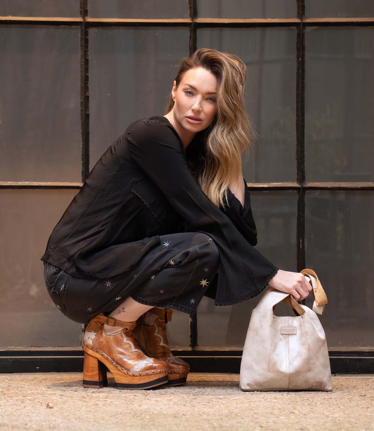 A woman squats in front of a window, wearing a black outfit with star details and brown vintage-inspired Bed|Stü Viretta platform boots featuring a handcrafted wooden platform, while holding a beige handbag.
