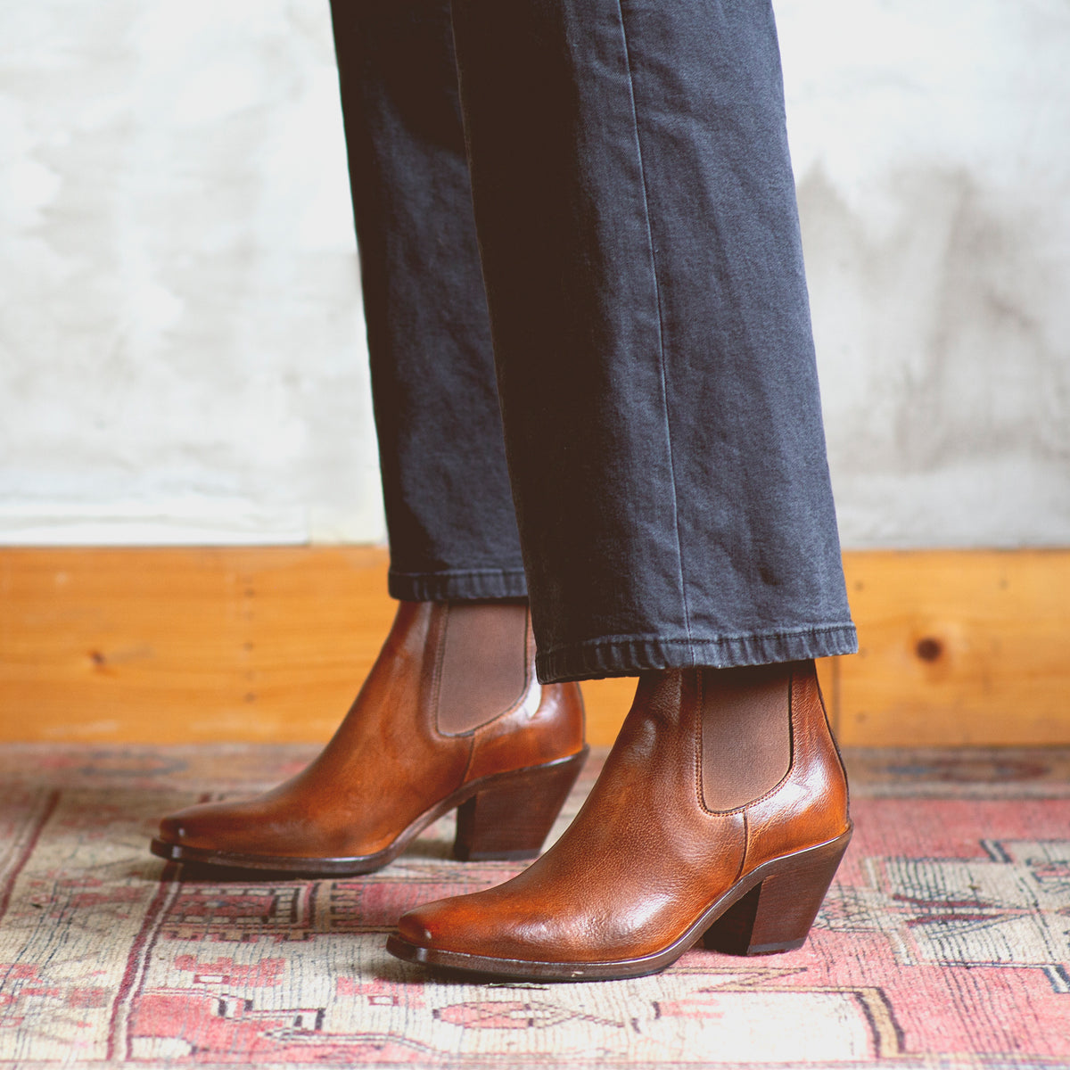 A person wearing the Bed|Stü Viscera brown full-grain leather heeled Chelsea boots and dark pants stands on a patterned rug, with a wooden baseboard and textured wall in the background.