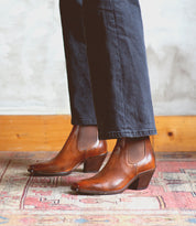 A person wearing the Bed|Stü Viscera brown full-grain leather heeled Chelsea boots and dark pants stands on a patterned rug, with a wooden baseboard and textured wall in the background.