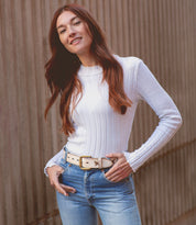 A woman with long reddish-brown hair, smiling with her hands on her hips, stands outdoors against a ridged wall wearing blue jeans, a white ribbed sweater, and the whimsical Bed|Stü Wander butterfly belt.