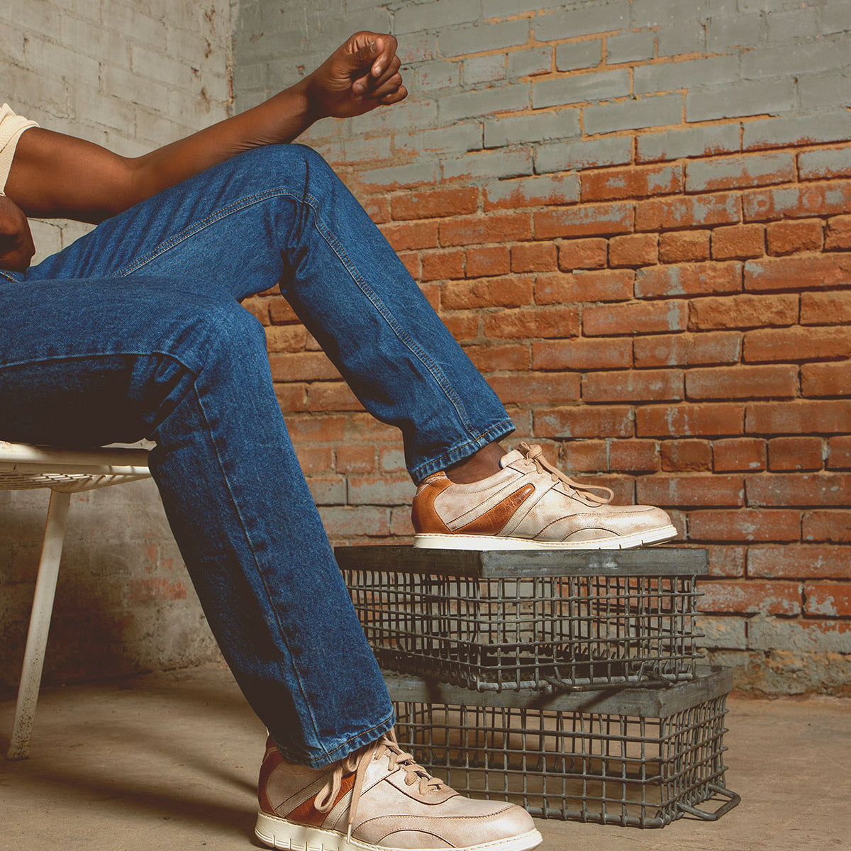 A person in jeans and the Bed|Stü Wardell layered leather sneakers with cushioned soles sits on a white chair beside stacked metal cages, set against a brick wall. Only their lower body and arm are visible, lending a modern edge to the scene.