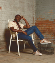 A man sits on a white metal chair in a brick-walled room, wearing a beige shirt, blue jeans, and the Bed|Stü Wardell layered leather sneakers with a cushioned sole, with two wire baskets stacked beside him.