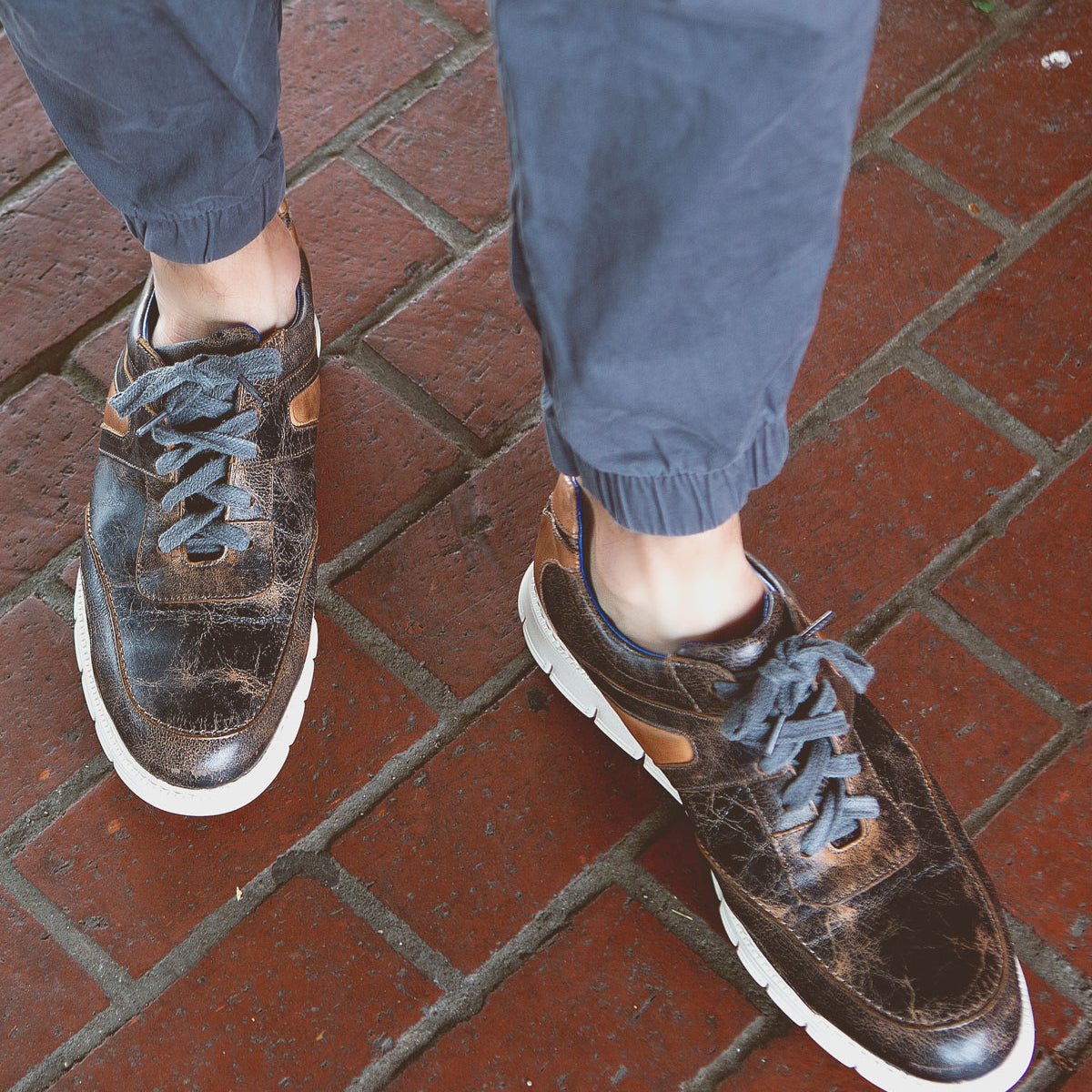 Wearing the Bed|Stü Wardell—an artfully worn brown layered leather sneaker with white cushioned soles and distinctive blue laces—a person stands on red brick pavement in cuffed blue pants for a modern edge.