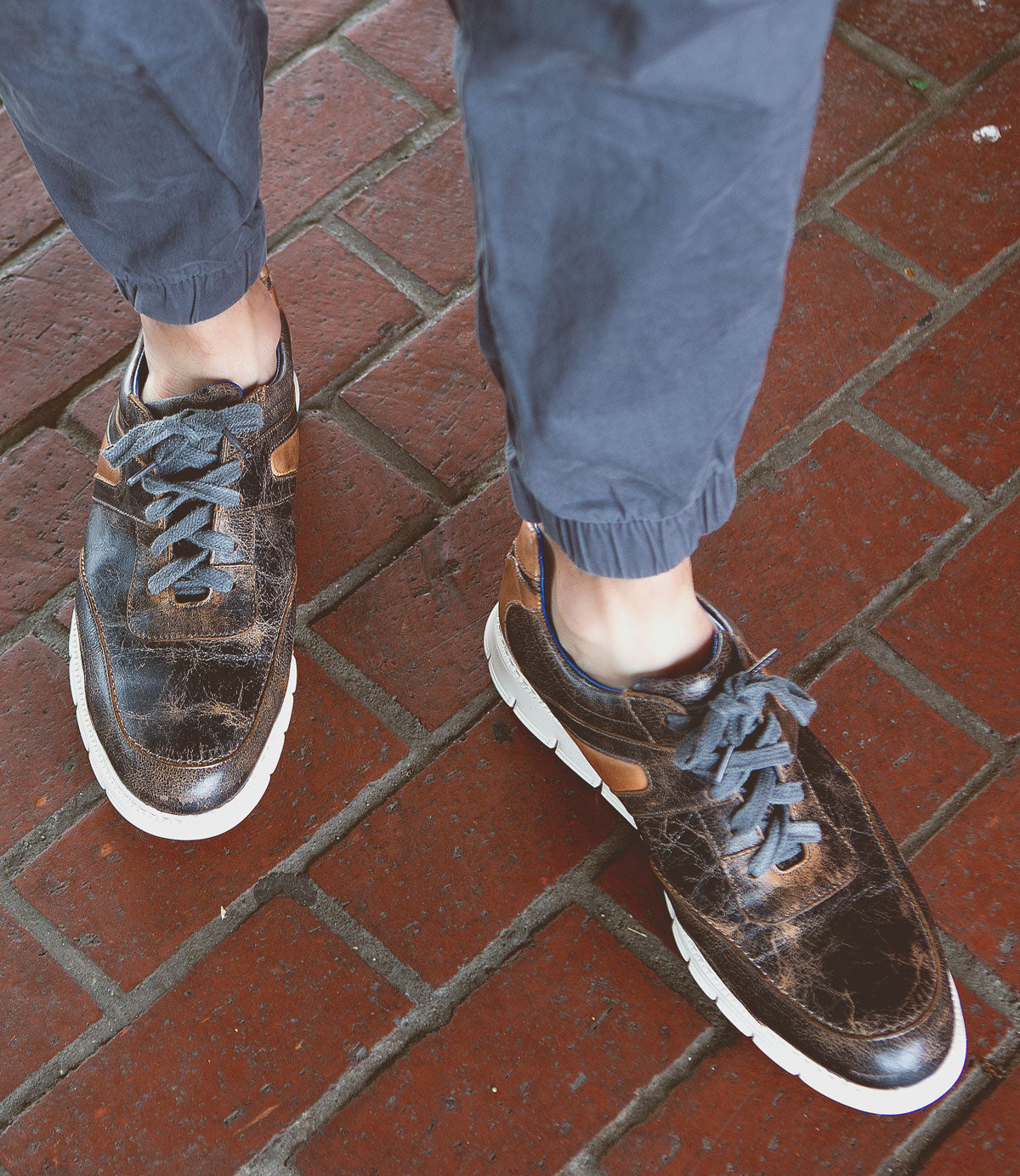 Wearing the Bed|Stü Wardell—an artfully worn brown layered leather sneaker with white cushioned soles and distinctive blue laces—a person stands on red brick pavement in cuffed blue pants for a modern edge.