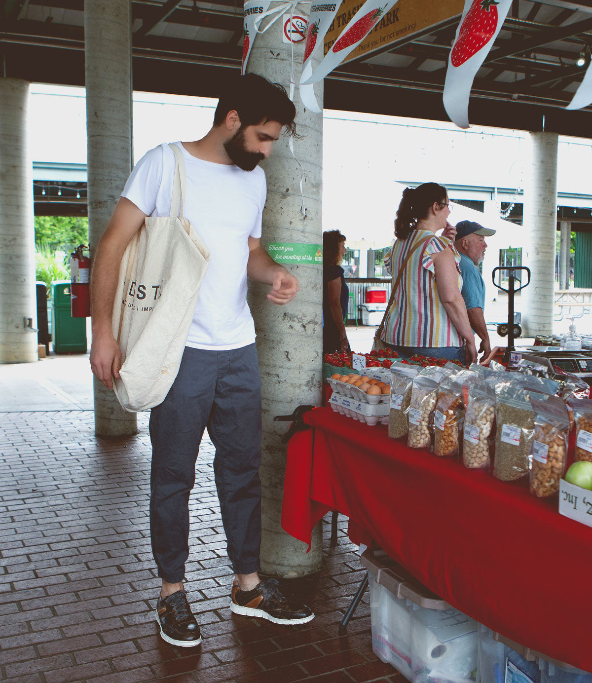 A man with a tote bag stands at a market stall with packaged goods on a red tablecloth, under a covered pavilion, his look finished with the Bed|Stü Wardell layered leather sneakers that add modern edge and cushioned sole comfort to his style.