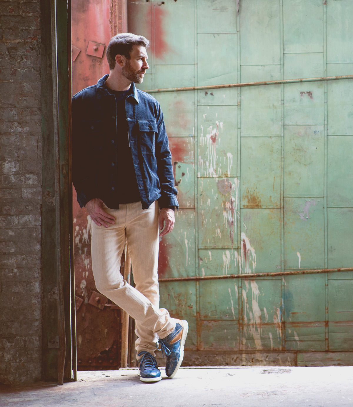 A man with a beard stands by an open, weathered green door in casual clothes, looking to the left with one foot crossed over the other, showcasing a modern edge in his layered Bed|Stü Wardell leather sneakers featuring cushioned soles.
