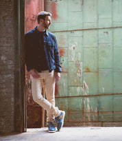A man with a beard stands by an open, weathered green door in casual clothes, looking to the left with one foot crossed over the other, showcasing a modern edge in his layered Bed|Stü Wardell leather sneakers featuring cushioned soles.