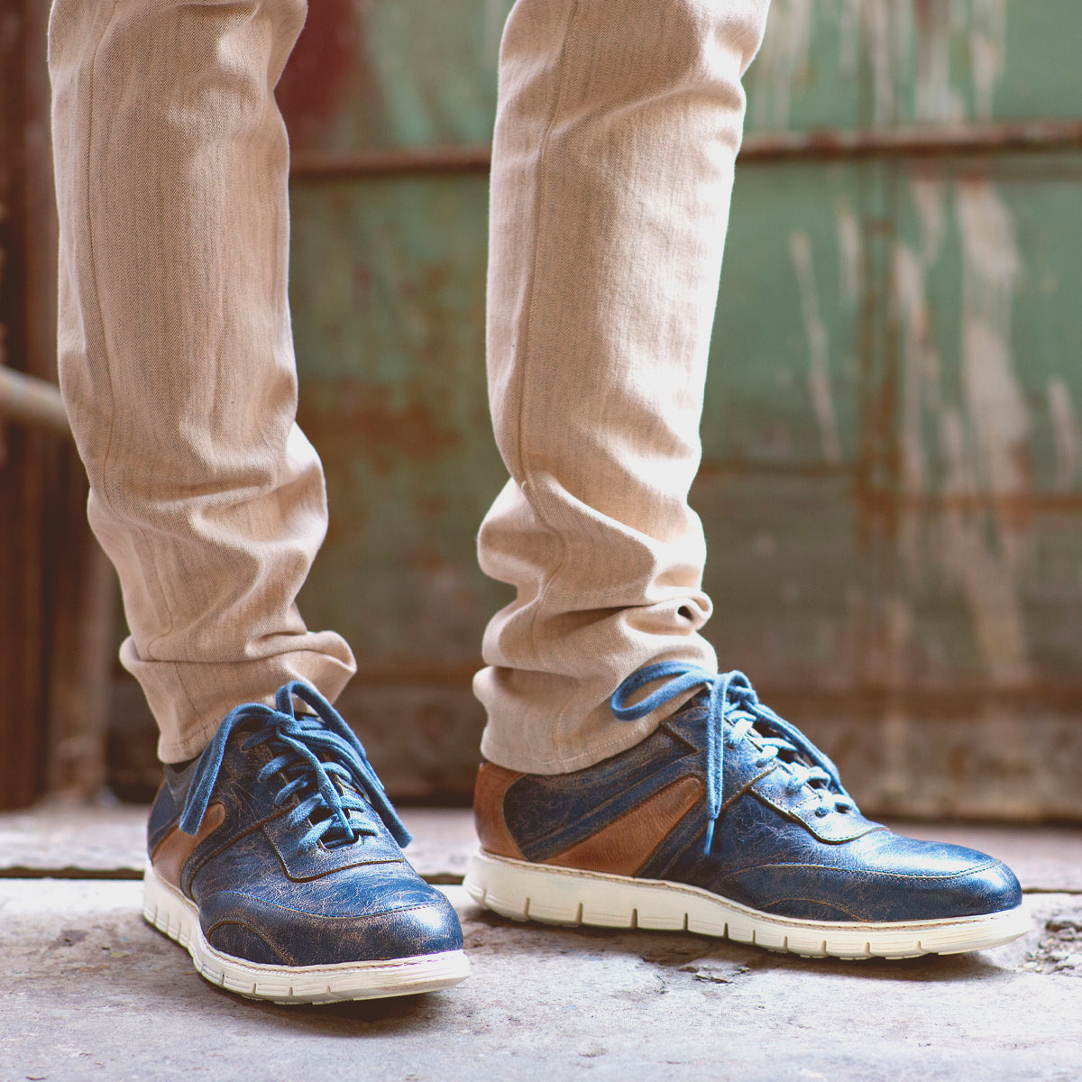 A person wearing beige pants and the Bed|Stü Wardell layered leather sneaker with a cushioned sole stands on a concrete surface in front of a weathered green metal door, bringing a modern edge to the scene.