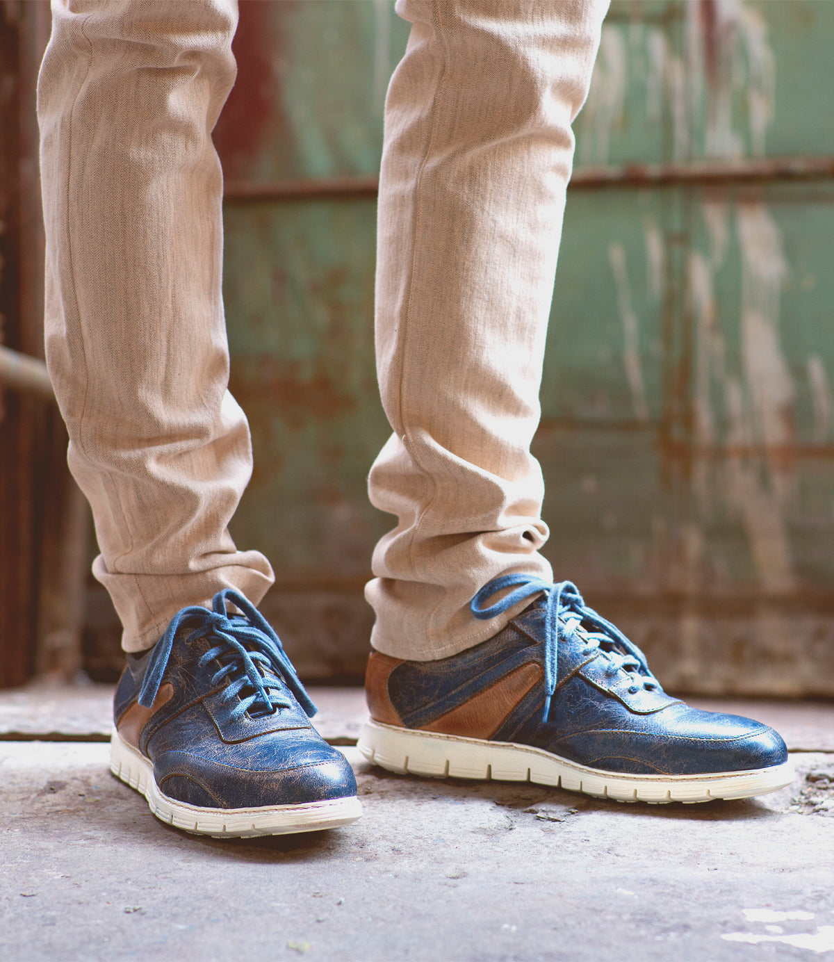 A person wearing beige pants and the Bed|Stü Wardell layered leather sneaker with a cushioned sole stands on a concrete surface in front of a weathered green metal door, bringing a modern edge to the scene.