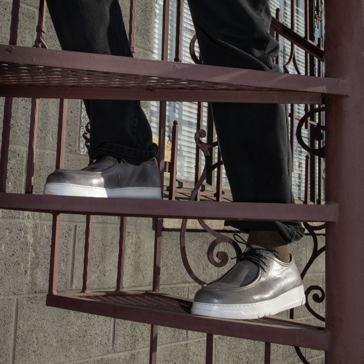 A person wearing the Bed|Stü Webmaster metallic silver moc-toe sneakers and black pants stands on a metal spiral staircase, with a brick wall and window in the background.