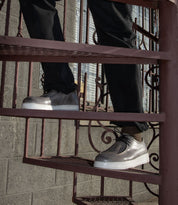 A person wearing the Bed|Stü Webmaster metallic silver moc-toe sneakers and black pants stands on a metal spiral staircase, with a brick wall and window in the background.