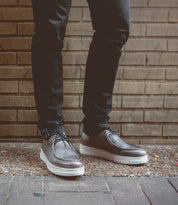A person wearing Bed|Stü’s Webmaster brown leather moc-toe sneakers with a white sole and lace-up design, paired with dark jeans, stands on a rough surface in front of a brick wall.