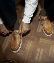 A person wearing the Bed|Stü Webmaster brown leather lace-up shoes paired with matching brown socks is tying their shoelaces while seated on a wooden parquet floor.
