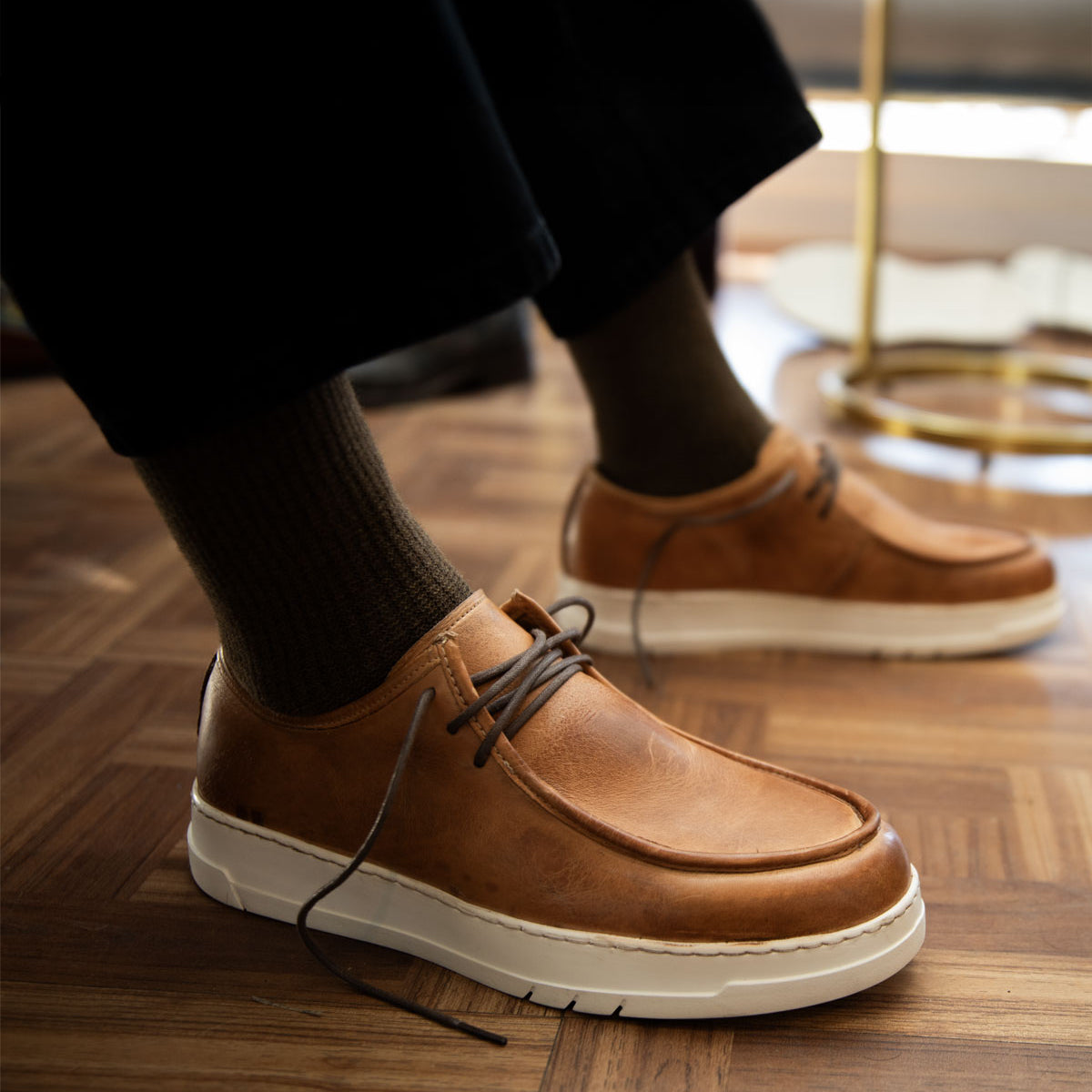 A person wearing the Bed|Stü Webmaster comfortable brown leather lace-up shoes with white soles and dark socks stands on a wooden floor.