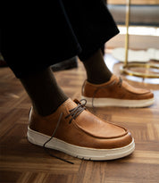 A person wearing the Bed|Stü Webmaster comfortable brown leather lace-up shoes with white soles and dark socks stands on a wooden floor.