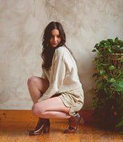 A woman with long dark hair crouches indoors on a wooden floor, wearing a cream sweater, light skirt, and stylish Bed|Stü Yell handmade leather boots, next to a green potted plant against a textured wall.