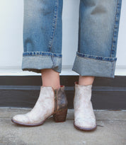 A person wearing the Bed|Stü Yell handmade leather ankle boots in a light, worn finish, featuring side zippers and block heels, paired with cuffed blue jeans and standing on concrete.