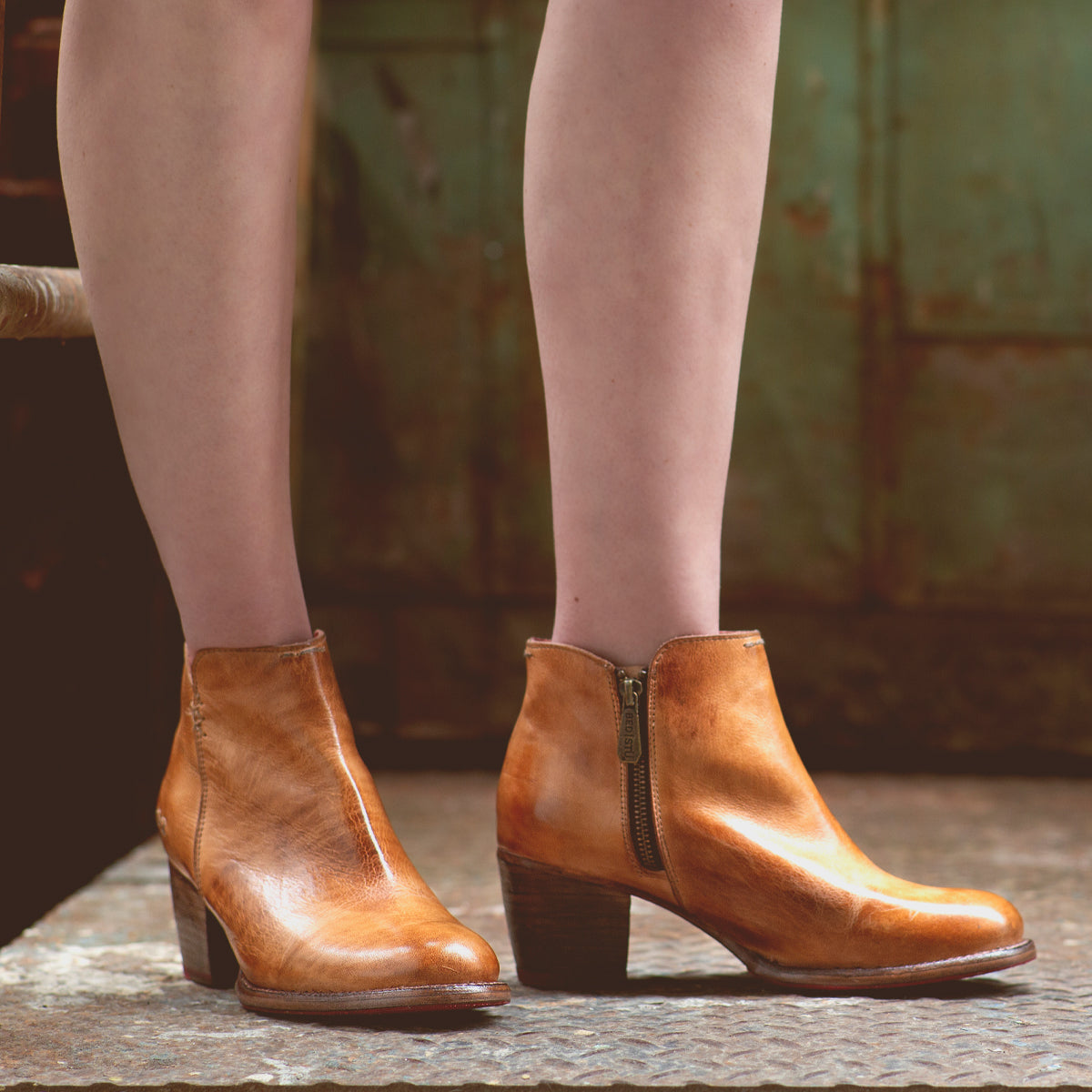 A person wearing the Bed|Stü "Yell" handmade leather ankle boots with zippers stands on a textured concrete surface, set against the backdrop of an aged green metal door.
