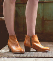 A person wearing the Bed|Stü "Yell" handmade leather ankle boots with zippers stands on a textured concrete surface, set against the backdrop of an aged green metal door.