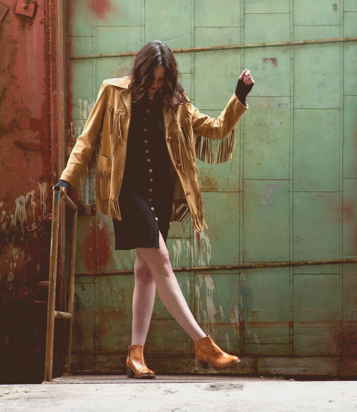 A woman in a tan fringed jacket and handmade Bed|Stü Yell leather boots stands on one leg near a metal railing, set against a green industrial door.