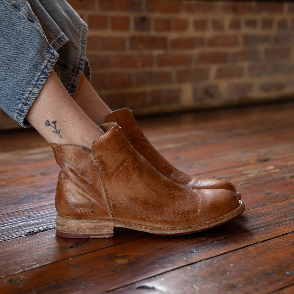 A person wearing the Yurisa handcrafted ankle boot by Bed|Stü, featuring a rich brown leather outsole, sits on a wooden floor with a brick wall behind them. Their jeans are cuffed to reveal a small flower tattoo on their ankle.