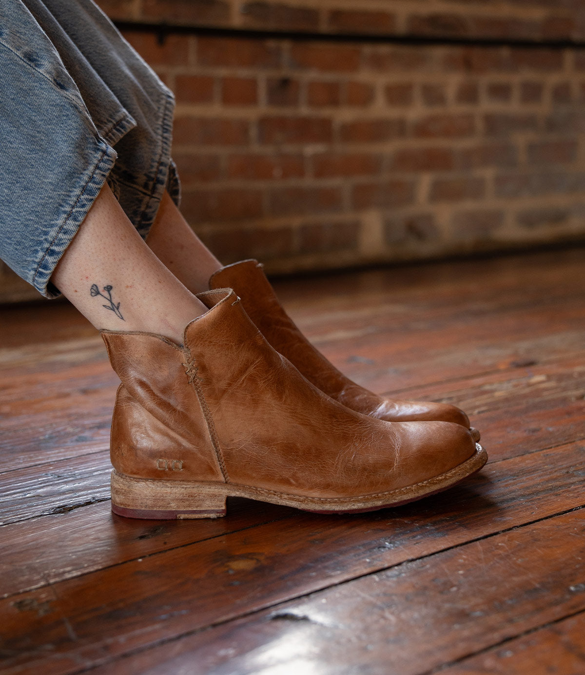 A person wearing the Yurisa handcrafted ankle boot by Bed|Stü, featuring a rich brown leather outsole, sits on a wooden floor with a brick wall behind them. Their jeans are cuffed to reveal a small flower tattoo on their ankle.