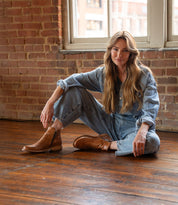 A woman in a denim outfit sits on a wooden floor in front of a brick wall and windows, looking at the camera with one leg bent and wearing stylish Bed|Stü Yurisa ankle boots featuring a sturdy leather outsole.