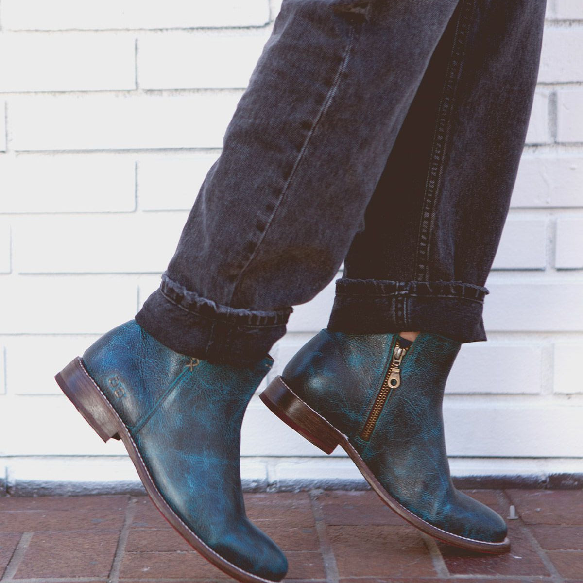 A person wearing rolled-up black jeans and Bed|Stü's Yurisa blue handcrafted leather ankle boots with side zippers, walking on a brick sidewalk in front of a white brick wall.