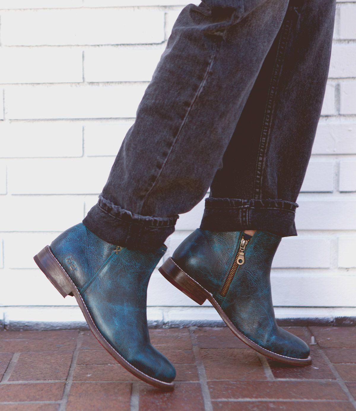 A person wearing rolled-up black jeans and Bed|Stü's Yurisa blue handcrafted leather ankle boots with side zippers, walking on a brick sidewalk in front of a white brick wall.
