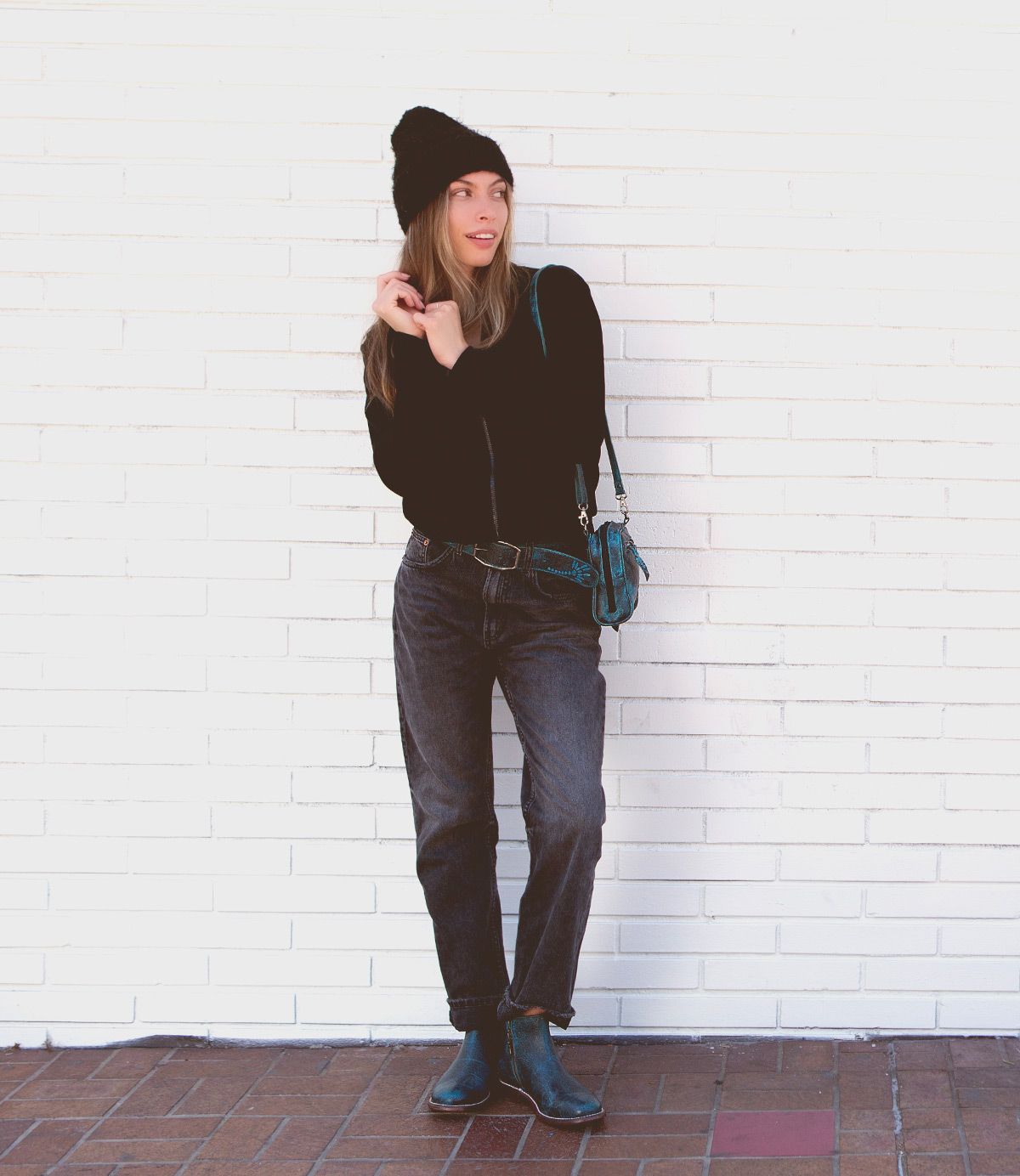 A woman stands against a white brick wall wearing a black beanie, black sweater, jeans, and ankle boots with a sleek leather outsole. She carries the Yurisa shoulder bag by Bed|Stü in teal and looks to the side with one hand near her face.