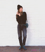 A woman stands against a white brick wall wearing a black beanie, black sweater, jeans, and ankle boots with a sleek leather outsole. She carries the Yurisa shoulder bag by Bed|Stü in teal and looks to the side with one hand near her face.
