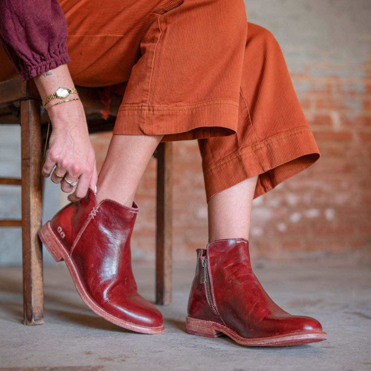 A person wearing brown corduroy pants and a maroon top sits on a wooden chair, showcasing the handcrafted Bed|Stü Yurisa ankle boot in red leather with convenient side zippers and a durable leather outsole.