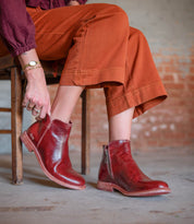 A person wearing brown corduroy pants and a maroon top sits on a wooden chair, showcasing the handcrafted Bed|Stü Yurisa ankle boot in red leather with convenient side zippers and a durable leather outsole.