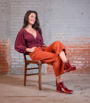 A woman with long brown hair sits on a wooden chair against a brick wall, wearing a maroon blouse, orange pants, and the stylish Yurisa red ankle boots by Bed|Stü featuring a leather outsole, smiling and looking to the side.