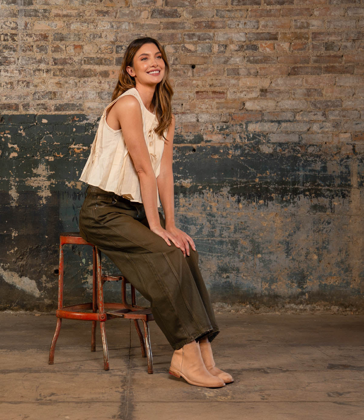 Smiling as she sits sideways on a red chair against a rustic brick wall, the woman wears a sleeveless beige top, dark green pants, and stylish Bed|Stü Yurisa tan ankle boots featuring leather outsoles.