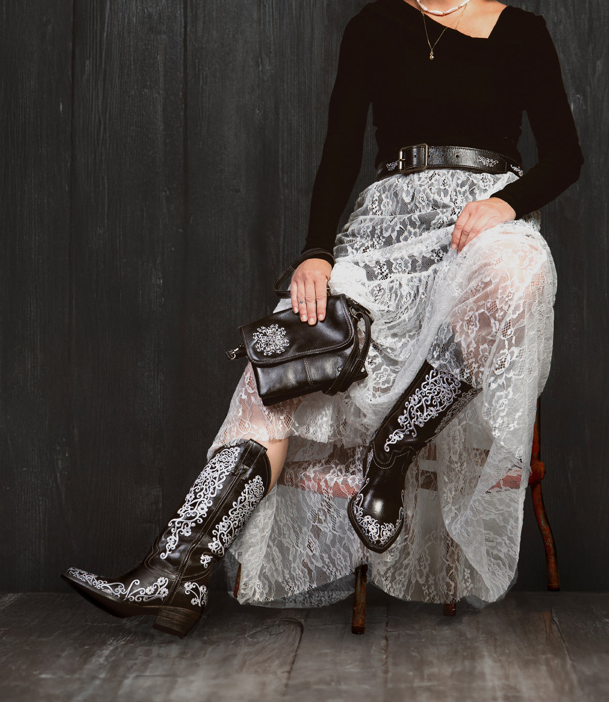 A person sits on a chair against a dark wooden background, wearing the Bed|Stü Ziggy JB black boots with white embroidery, paired with a black top and lace skirt, and holding a sleek clutch.