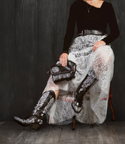 A person sits on a chair against a dark wooden background, wearing the Bed|Stü Ziggy JB black boots with white embroidery, paired with a black top and lace skirt, and holding a sleek clutch.