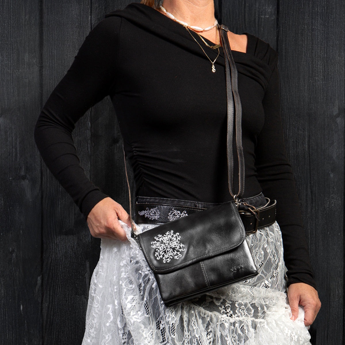 A woman wearing a black top and a white lace skirt holds the Bed|Stü Ziggy JB black leather crossbody bag, featuring a silver floral embellishment crafted from organic cotton accents, set against a dark wooden background.