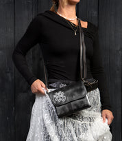 A woman wearing a black top and a white lace skirt holds the Bed|Stü Ziggy JB black leather crossbody bag, featuring a silver floral embellishment crafted from organic cotton accents, set against a dark wooden background.